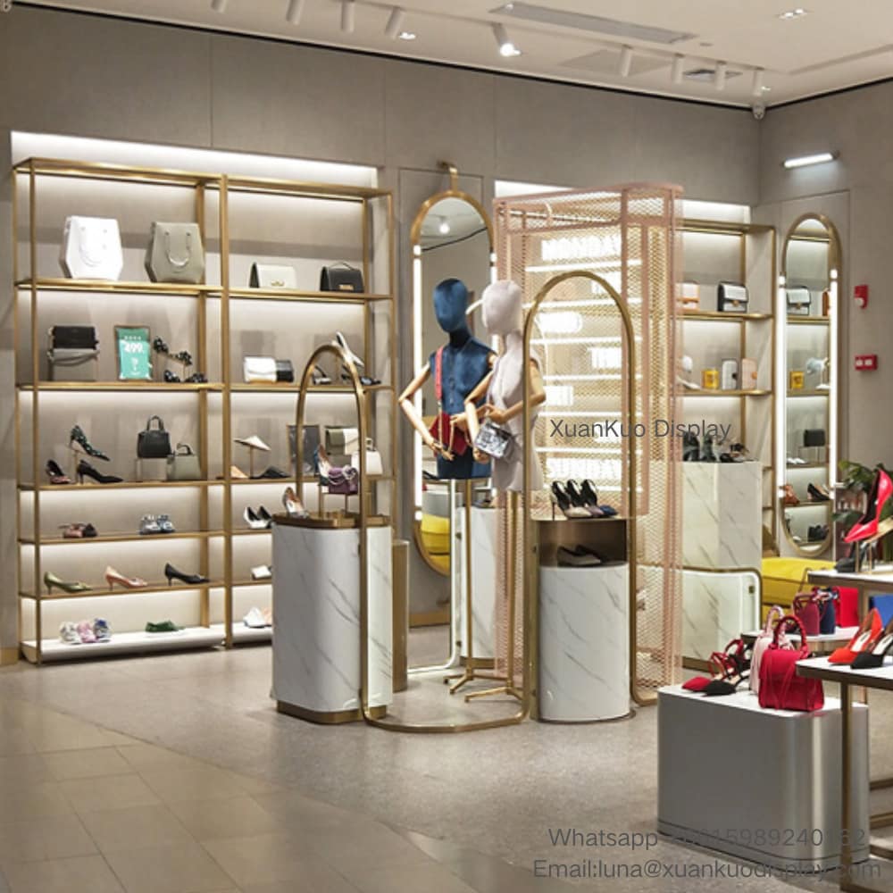 One-stop display furniture solution for women’s shoe and handbag boutique. Surrounding wall adopts gold metal frame multi-layer backlit shelves, neatly arranging high heels, sandals and leather handbags. Central area equipped with white marble pattern cylindrical display plinths and arched gold mirror mannequin stands for outfit showcase. Perforated gold slat wall serves as space divider and brand decorative zone, bright elegant tone forms high-quality shopping atmosphere, suitable for shopping mall leather goods retail stores.